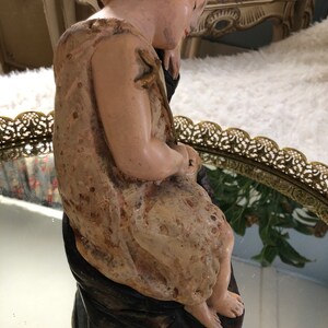 May include: A vintage figurine of a child sitting with a contemplative expression. The figurine is made of painted plaster and has a textured surface. The child is wearing a simple garment and has a delicate, almost ethereal quality.