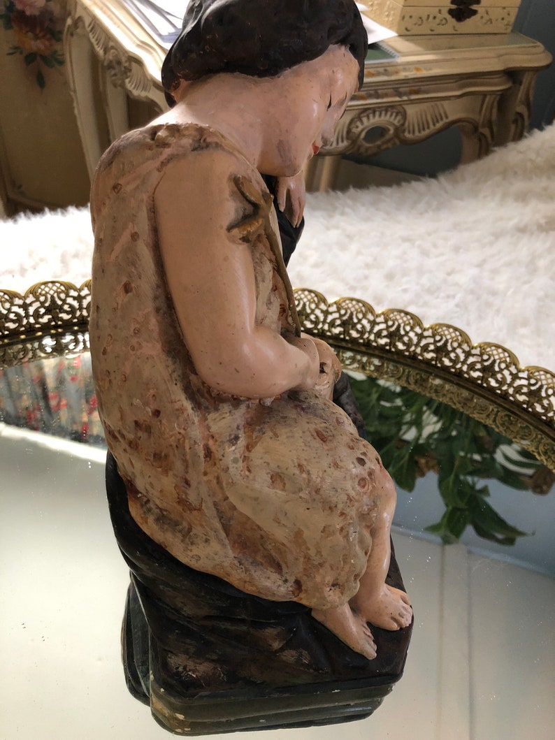 May include: A vintage figurine of a child sitting with a contemplative expression. The figurine is made of painted plaster and has a textured surface. The child is wearing a simple garment and has a delicate, almost ethereal quality.
