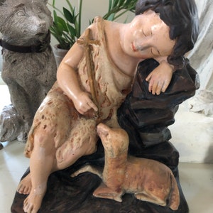 May include: A vintage figurine of a sleeping boy with a lamb, made of painted plaster. The boy is wearing a simple robe and holding a staff with a cross. The figurine is set on a brown base.