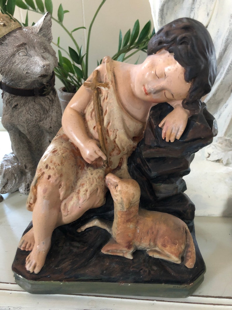 May include: A vintage figurine of a sleeping boy with a lamb, made of painted plaster. The boy is wearing a simple robe and holding a staff with a cross. The figurine is set on a brown base.