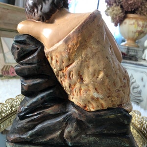 May include: A vintage plaster statue of a young person sitting on a rock. The statue is painted in shades of brown, tan, and white. The figure is wearing a simple garment and has a relaxed pose.