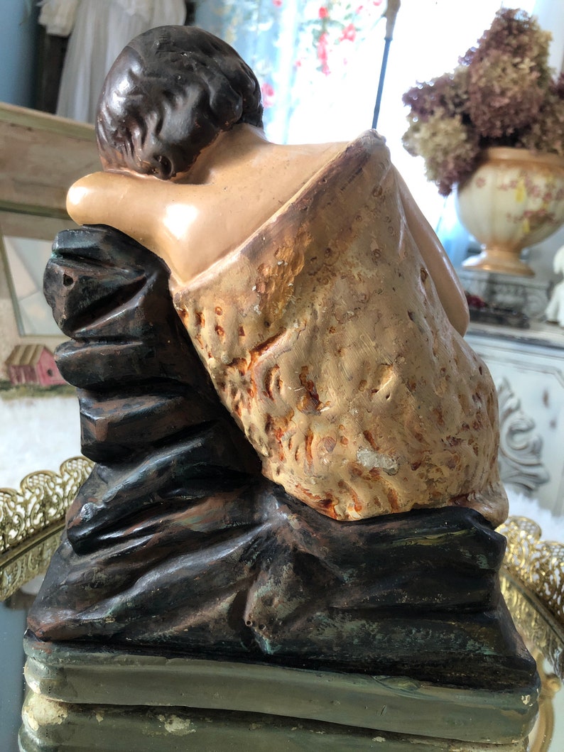 May include: A vintage plaster statue of a young person sitting on a rock. The statue is painted in shades of brown, tan, and white. The figure is wearing a simple garment and has a relaxed pose.