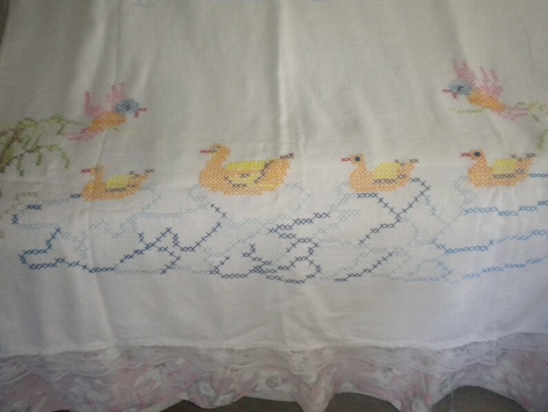 May include: White cotton tablecloth with embroidered ducks and birds in yellow, blue, pink, and green. The tablecloth has a lace trim.