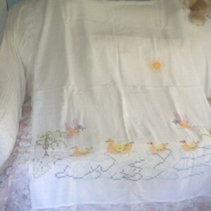 May include: White embroidered tablecloth with a sun, clouds, birds, and ducks. The tablecloth has a lace trim.