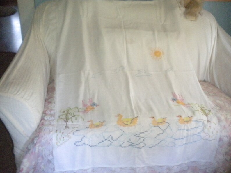 May include: White embroidered tablecloth with a sun, clouds, birds, and ducks. The tablecloth has a lace trim.