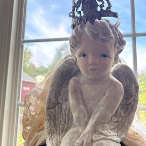 Pode incluir: Uma estatueta de anjo sentado com asas detalhadas e uma coroa decorativa. O anjo é pintado em um tom neutro, com um acabamento texturizado. O fundo inclui uma janela com vista para o céu e árvores.