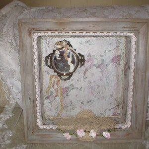 May include: A framed shadow box with a floral patterned background. The box is decorated with lace, a small mirror, and a few artificial flowers.