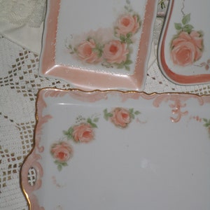 Könnte beinhalten: Drei weiße Porzellanserviertabletts mit rosafarbenen Blumendesigns. Die Tabletts haben einen gewellten Rand und einen goldenen Rand. Die Blumen sind in Rosatönen und Weiß bemalt.