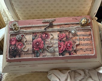 May include: A vintage-style wooden box with a distressed pink and white paint finish. The box is decorated with a floral design of pink roses, musical notes, and rhinestone accents. A key and decorative brooches enhance the antique look.
