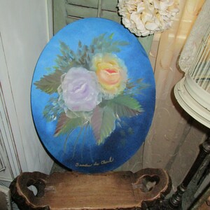 May include: Oval painting with a bouquet of roses in yellow, lavender, and peach hues, set against a deep blue backdrop. The artwork is signed "Sandra du Chard". The painting is displayed on a wooden bench.