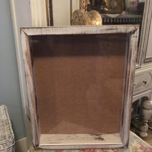 May include: A white wooden shadow box with a distressed finish. The box has a clear glass front and a brown cardboard back.