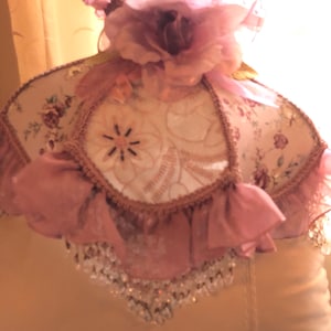 May include: A delicate lampshade with a pink floral design and a ruffled trim. The lampshade is adorned with a large pink flower.