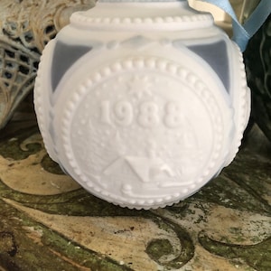 May include: White porcelain Christmas bauble with a blue ribbon. The bauble has a raised design of a house and the year 1988.