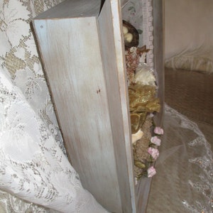 May include: A light blue wooden shadow box with a distressed finish. The box is filled with various decorative items, including dried flowers, lace, and a small bird's nest.