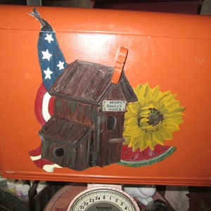 May include: Orange painted wooden box with a hand-painted design featuring a house, a sunflower, a watermelon, and an American flag. The house has a sign that reads "Home Sweet Home".