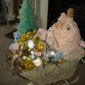 May include: A white sleigh filled with a variety of Christmas ornaments, including gold, green, and pink ornaments. A pink doll wearing a white lace dress sits in the sleigh.