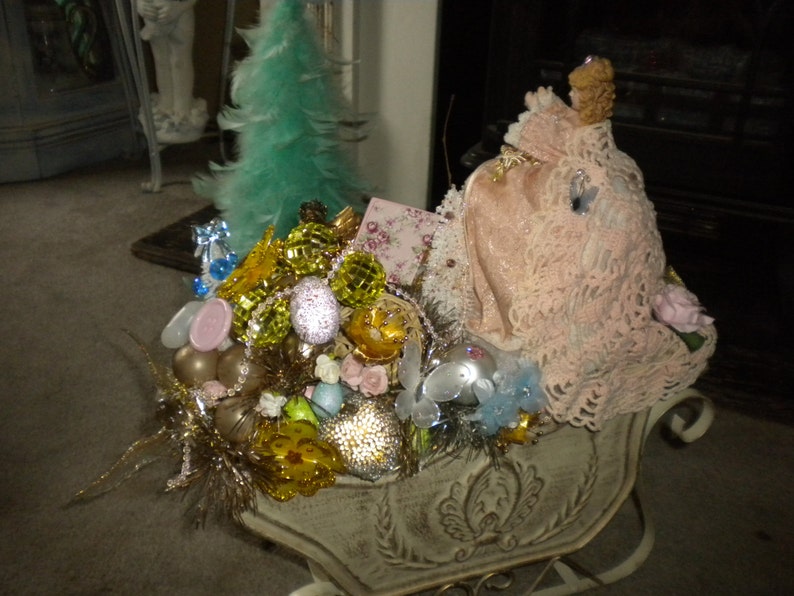May include: A white sleigh filled with a variety of Christmas ornaments, including gold, green, and pink ornaments. A pink doll wearing a white lace dress sits in the sleigh.