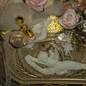May include: A white ceramic angel figurine with gold accents and a white lace trim. The angel is holding a small white bag with gold trim. The figurine is surrounded by gold and white decorative elements.