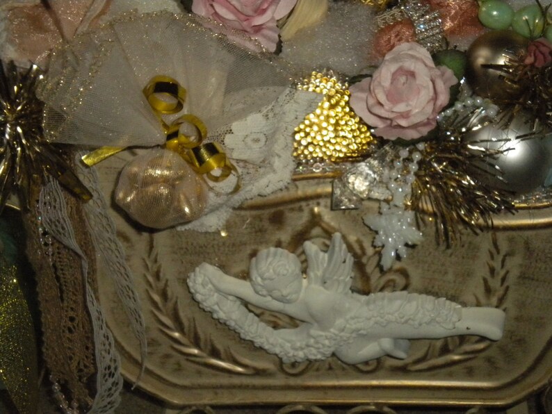 May include: A white ceramic angel figurine with gold accents and a white lace trim. The angel is holding a small white bag with gold trim. The figurine is surrounded by gold and white decorative elements.