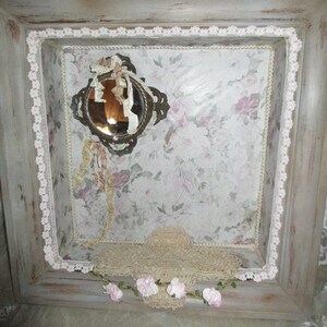 May include: A gray wooden frame with a white lace border. Inside the frame is a floral patterned paper with a small antique mirror and a lace doily. The frame is decorated with a garland of pink flowers.