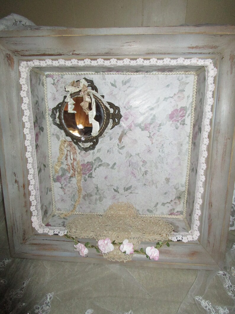 May include: A gray wooden frame with a white lace border. Inside the frame is a floral patterned paper with a small antique mirror and a lace doily. The frame is decorated with a garland of pink flowers.