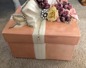 May include: A ceramic gift box shaped like a present with a white ribbon and a pink bow. The bow is decorated with pink and white flowers and rhinestones.