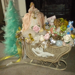 May include: A white sleigh filled with a variety of Christmas decorations, including a small white angel, a miniature feather Christmas tree, a porcelain doll, and a variety of ornaments. The sleigh is decorated with gold ribbon and white lace.