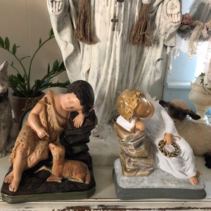 May include: Two vintage religious figurines, one depicting a sleeping angel with a crown of thorns and the other a shepherd boy with a lamb. Both are painted in muted tones of white, brown, and gold.