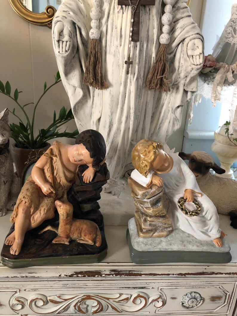 May include: Two vintage religious figurines, one depicting a sleeping angel with a crown of thorns and the other a shepherd boy with a lamb. Both are painted in muted tones of white, brown, and gold.
