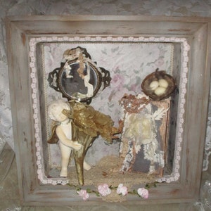 Peut inclure: Une boîte à ombre encadrée avec une esthétique vintage. Elle présente une figurine de chérubin en porcelaine tenant une coquille dorée, un petit miroir à motif floral, un nid d'oiseau avec trois œufs et un livre avec des embellissements en dentelle. La boîte est décorée d'une bordure en dentelle rose et d'une guirlande de fleurs roses.