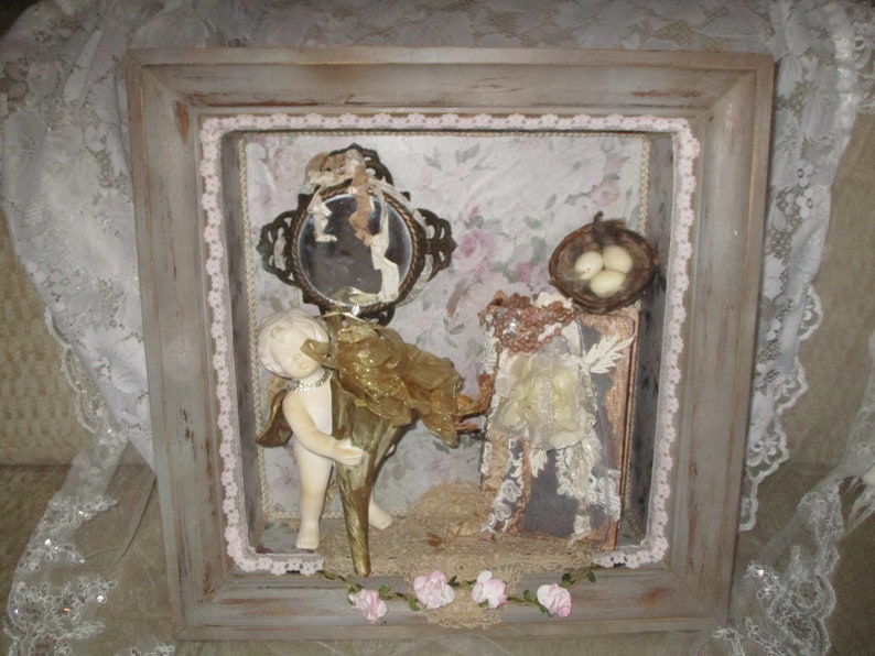 May include: A framed shadow box with a vintage aesthetic. It features a porcelain cherub figurine holding a golden shell, a small mirror with a floral design, a bird's nest with three eggs, and a book with lace embellishments. The box is decorated with a pink lace trim and a garland of pink flowers.