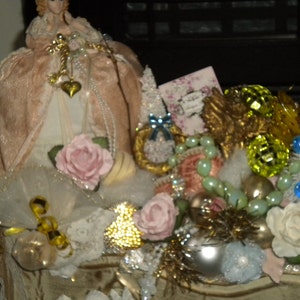 May include: A vintage-inspired Christmas decoration featuring a porcelain doll wearing a pink dress, surrounded by a variety of ornaments, including a gold angel, a pink rose, and a green and blue beaded garland.