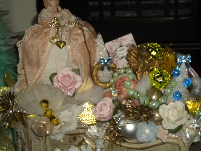 May include: A vintage-inspired Christmas decoration featuring a porcelain doll wearing a pink dress, surrounded by a variety of ornaments, including a gold angel, a pink rose, and a green and blue beaded garland.