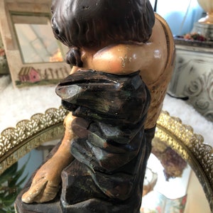 May include: A vintage figurine of a person with brown hair and a brown and green robe sitting on a rock. The figurine is made of plaster and has a distressed finish.