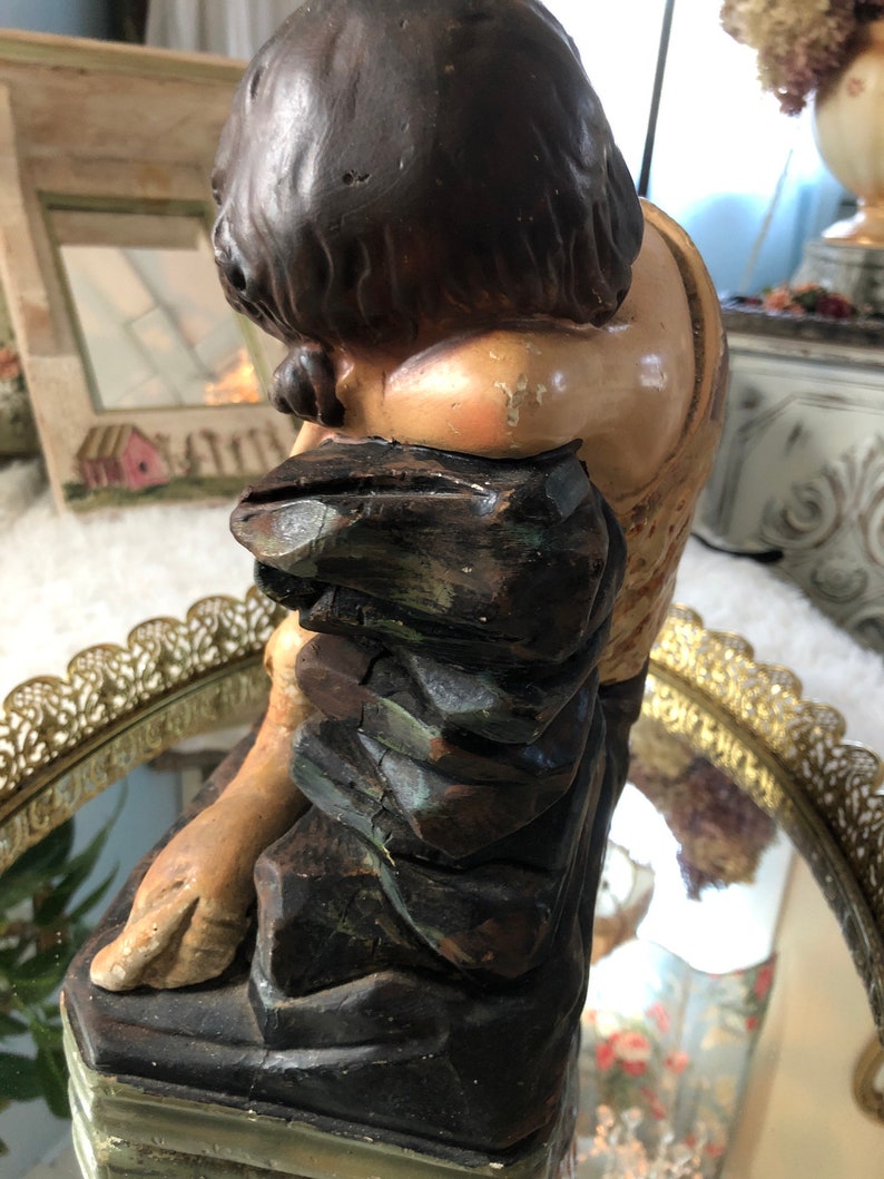 May include: A vintage figurine of a person with brown hair and a brown and green robe sitting on a rock. The figurine is made of plaster and has a distressed finish.
