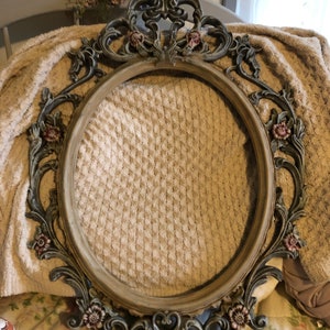 Puede incluir: Un marco de fotos ovalado adornado con un acabado desgastado y envejecido. El marco está hecho de metal y tiene un diseño floral decorativo en un esquema de color verde oscuro y marrón.
