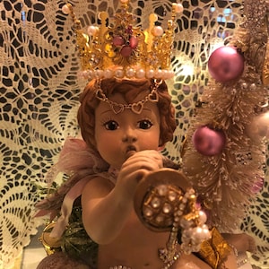 Puede incluir: Una figurita de querubín de porcelana que lleva una corona dorada con detalles en rosa y blanco, sosteniendo una trompeta dorada con pedrería. El querubín está sentado sobre una base con un borla dorada y pedrería.