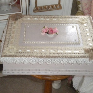 May include: A light purple wooden box with a lace and ribbon trim. The lid is decorated with a pearl border and a small cluster of pink flowers.