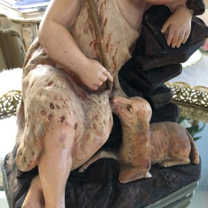 May include: A vintage figurine of a sleeping child holding a staff with a cross, with a small dog at their feet. The figurine is made of painted plaster and has a rustic, aged appearance.