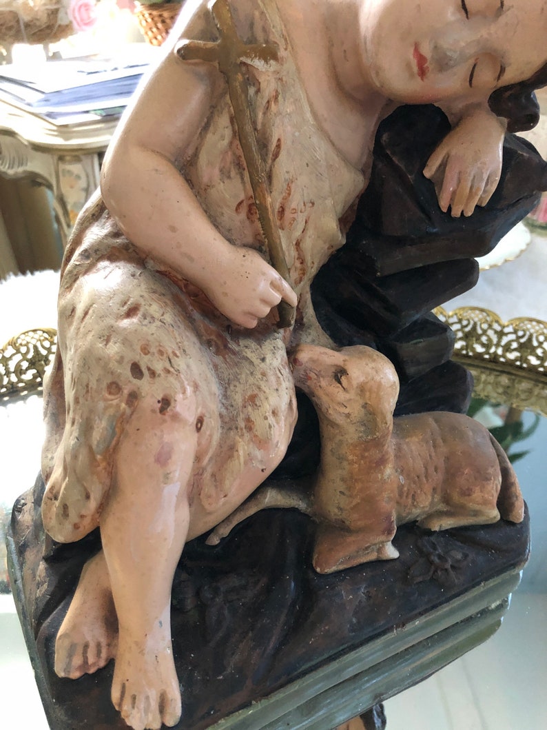 May include: A vintage figurine of a sleeping child holding a staff with a cross, with a small dog at their feet. The figurine is made of painted plaster and has a rustic, aged appearance.