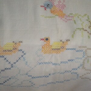 May include: A white fabric with a cross-stitch design of a yellow duck, a blue and yellow duck, and a green tree. The ducks are swimming in blue water and the tree is on a brown island.