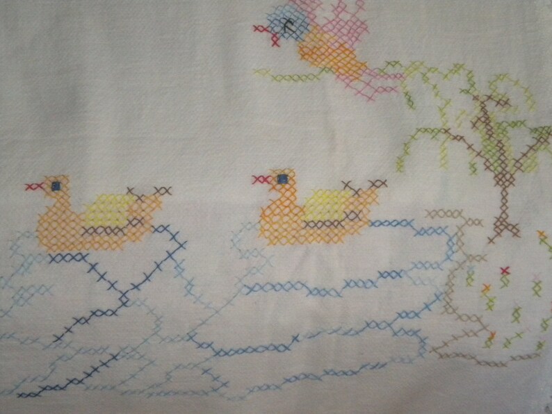 May include: A white fabric with a cross-stitch design of a yellow duck, a blue and yellow duck, and a green tree. The ducks are swimming in blue water and the tree is on a brown island.