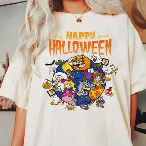 Könnte beinhalten: Cremefarbenes T-Shirt mit einer farbenfrohen Halloween-Grafik. Das Design zeigt verschiedene Zeichentrickfiguren in Halloween-Kostümen, mit den Worten "HAPPY HALLOWEEN" in Orange über den Figuren.