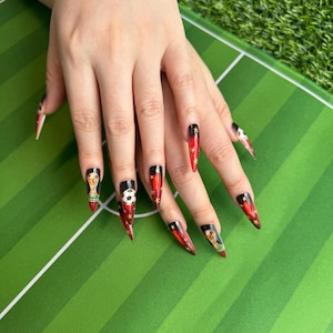 May include: Long, pointed fingernails with a soccer theme. The nails feature a black base with red and gold accents, including stars, soccer balls, and a trophy. The nails are on a green surface.
