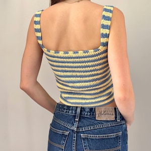 K&ouml;nnte beinhalten: Ein &auml;rmelloses H&auml;keloberteil mit horizontalen Streifen in Hellblau und Blassgelb. Das Kleidungsst&uuml;ck hat einen eckigen Ausschnitt und breite Tr&auml;ger und wird mit einer dunkelblauen Jeans kombiniert, die einen braunen Lederaufn&auml;her am Bund hat.