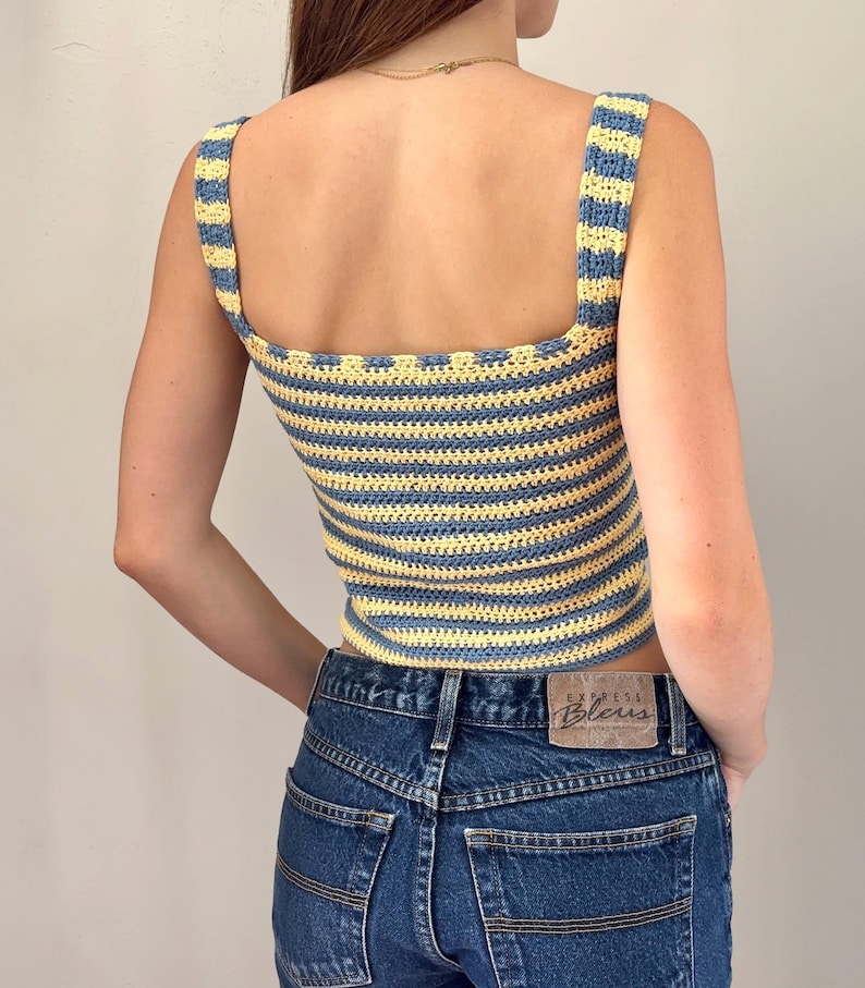 K&ouml;nnte beinhalten: Ein &auml;rmelloses H&auml;keloberteil mit horizontalen Streifen in Hellblau und Blassgelb. Das Kleidungsst&uuml;ck hat einen eckigen Ausschnitt und breite Tr&auml;ger und wird mit einer dunkelblauen Jeans kombiniert, die einen braunen Lederaufn&auml;her am Bund hat.
