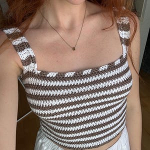 K&ouml;nnte beinhalten: Ein &auml;rmelloses H&auml;kel-Bustier mit horizontalen Streifen in abwechselndem Braun und Wei&szlig;. Das Kleidungsst&uuml;ck hat einen eckigen Ausschnitt und breite Tr&auml;ger, gefertigt mit einem durchgehend gleichm&auml;&szlig;igen, strukturierten Maschenmuster.