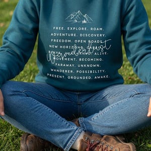Könnte beinhalten: Türkisfarbenes Sweatshirt mit Rundhalsausschnitt, weißem Text und einer Berg-Grafik. Der Text enthält Sätze wie "Free. Explore. Roam." und "Adventure. Discovery." Die Person trägt eine blaue Jeans und braune Wanderstiefel.