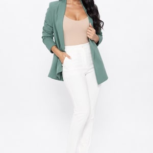 May include: A woman wearing a sage green blazer, a beige top, and white pants. The blazer has long sleeves and a draped collar. The pants are fitted. The woman is also wearing beige high-heeled sandals. The background is white.
