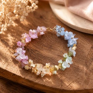 May include: A floral bracelet in a spectrum of colours, from pink to green, with gold-coloured butterfly details. The bracelet is displayed on a wooden surface. The bracelet's design incorporates a variety of small, flower-shaped beads.
